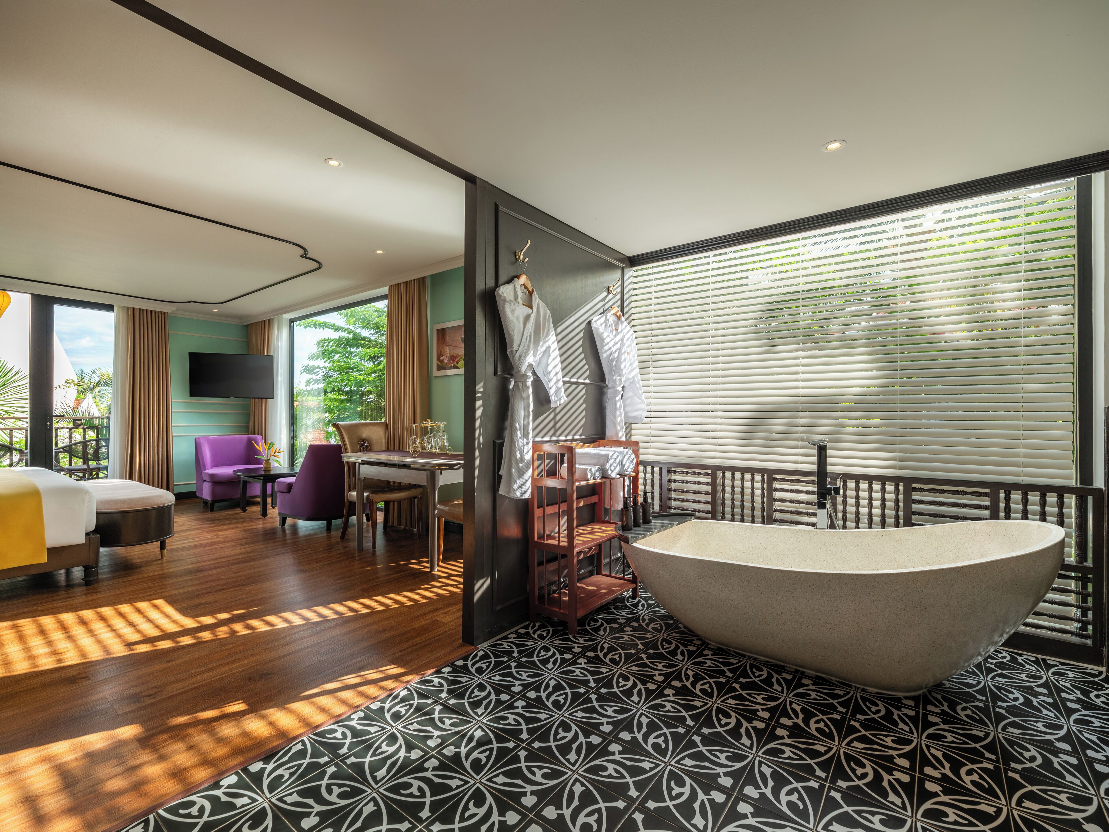 Una habitación de hotel moderna con una bañera grande, una zona de comedor con una mesa y sillas, un sofá y un balcón con vistas a zonas verdes.