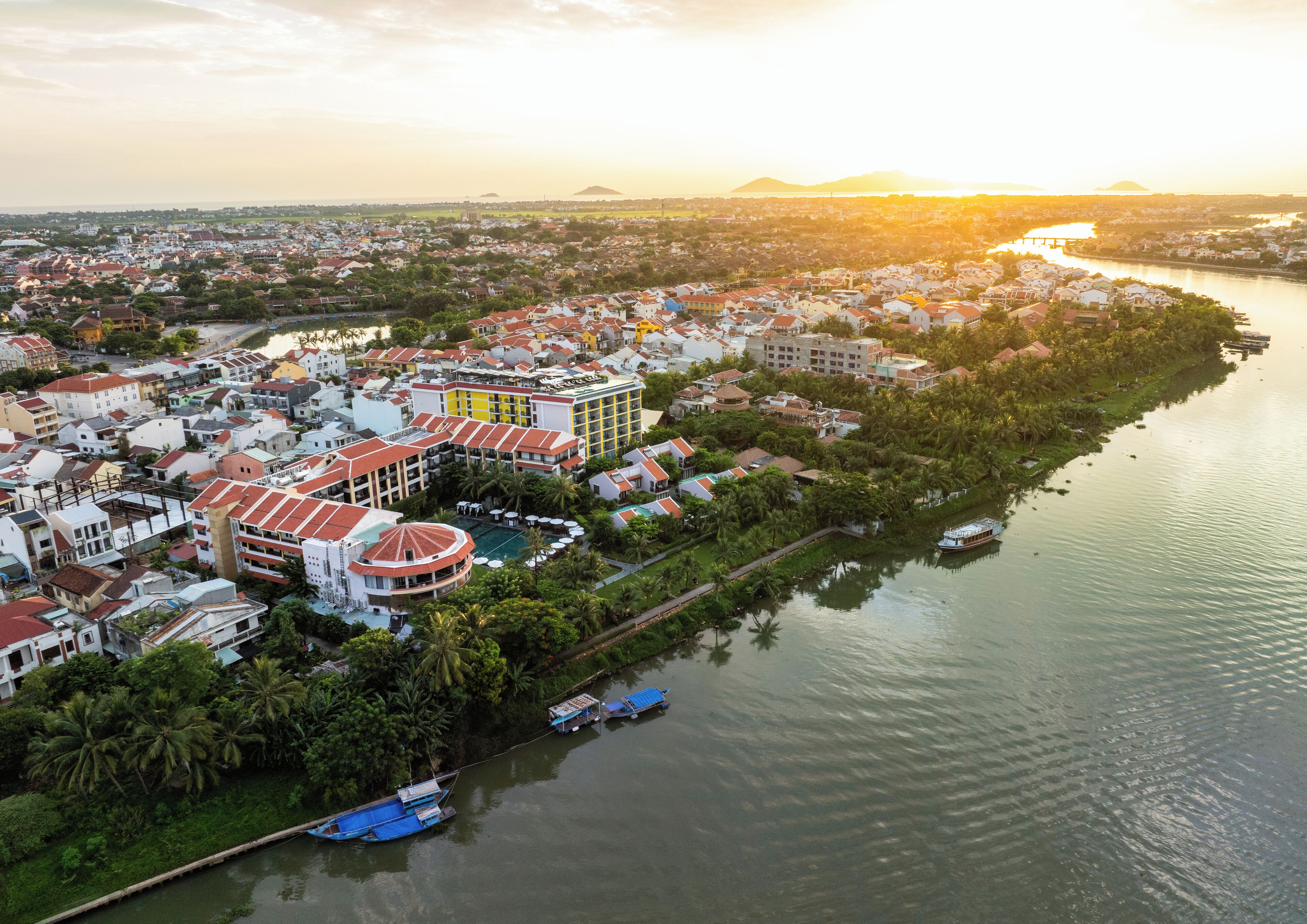 An aerial view of a coastal town with a prominent hotel featuring a circular pool and multiple buildings with red roofs.