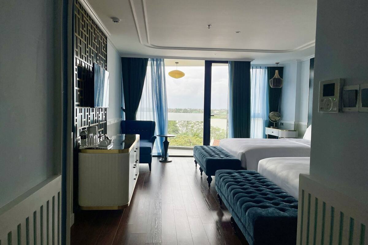 A hotel room with a bed, a bench, a small table, a dresser, and a view of the outdoors.