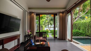 A modern living room with a flat-screen TV, a coffee table with fruit and a vase, a sofa, and a view of a lush garden and pool. - Bel Marina Hoi An Resort (Da Nang)