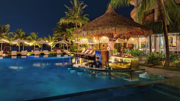 A nighttime pool area with lounge chairs, a bar, and thatched umbrellas. - Bel Marina Hoi An Resort (Da Nang)
