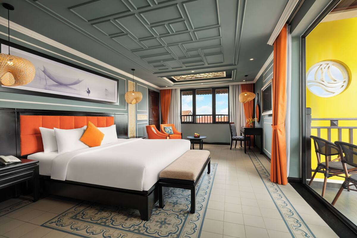 A hotel room with a large bed, a desk, a chair, a small table, and a balcony with chairs.