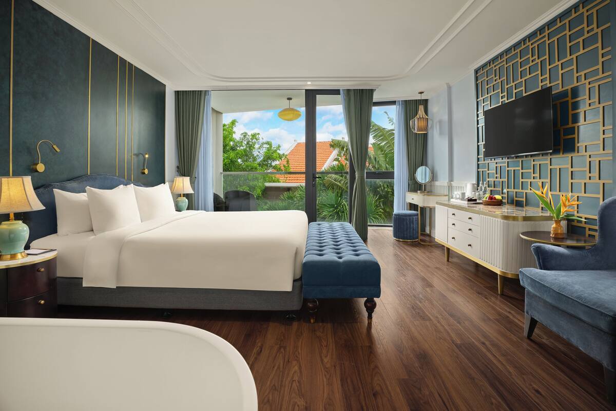 A hotel room with a large bed, a sitting area, a vanity, and a television.