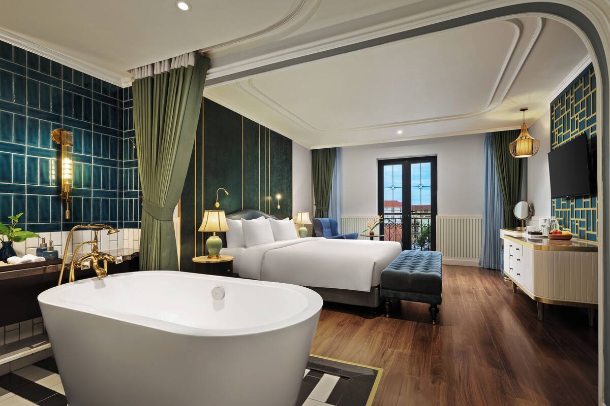 A luxurious hotel room with a large bathtub, a double bed, a sitting area, and a balcony with a view.