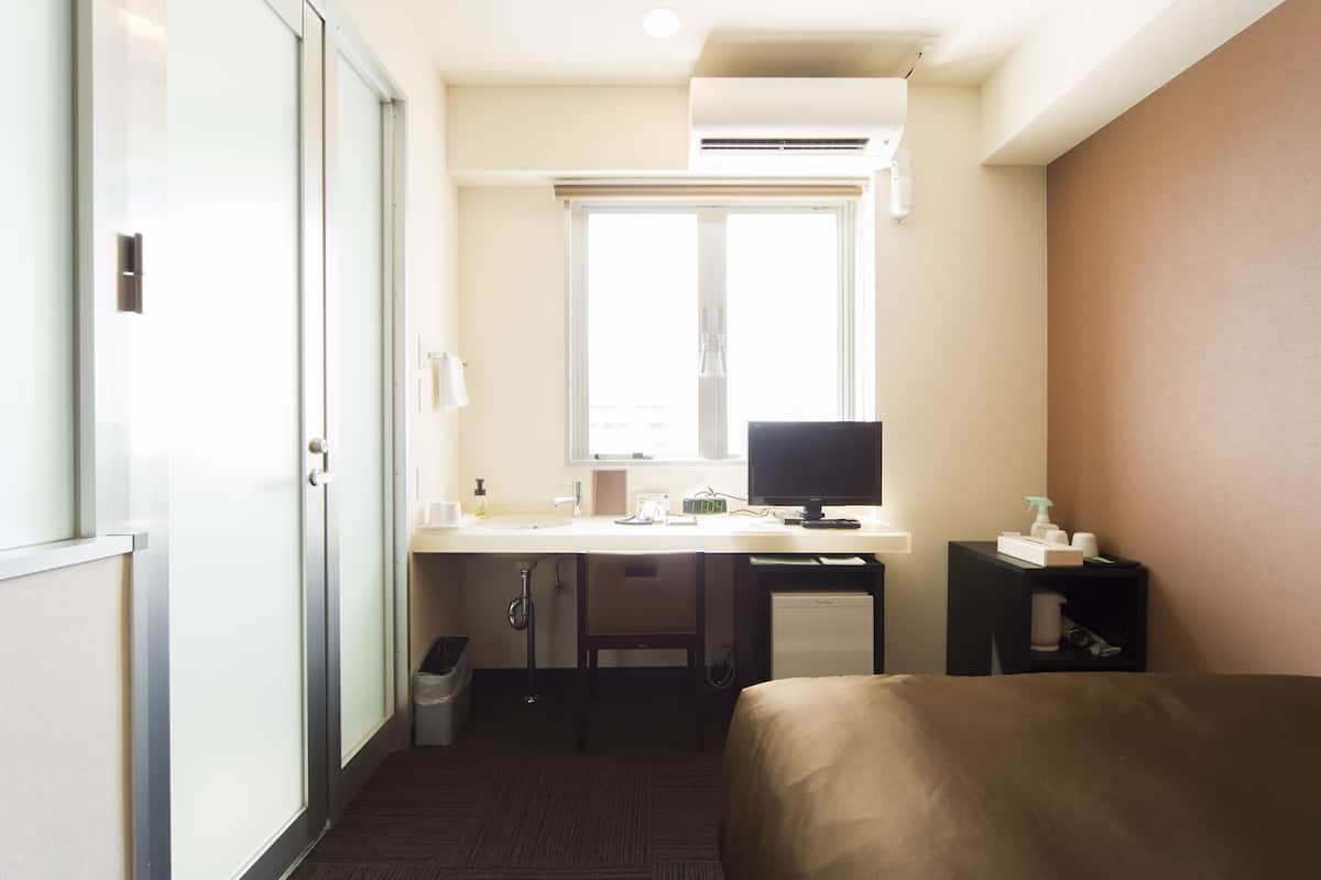 A hotel room with a bed, a desk with a computer, a chair, a small refrigerator, a trash bin, and a window with blinds.