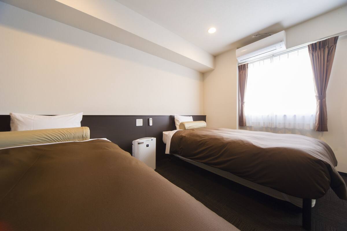 A hotel room with two beds, a bedside table, a lamp, and a window with curtains.