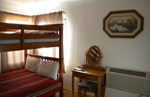 Una cama litera con una colcha a rayas rojas y blancas, un escritorio de madera con un espejo, y un cuadro en la pared.