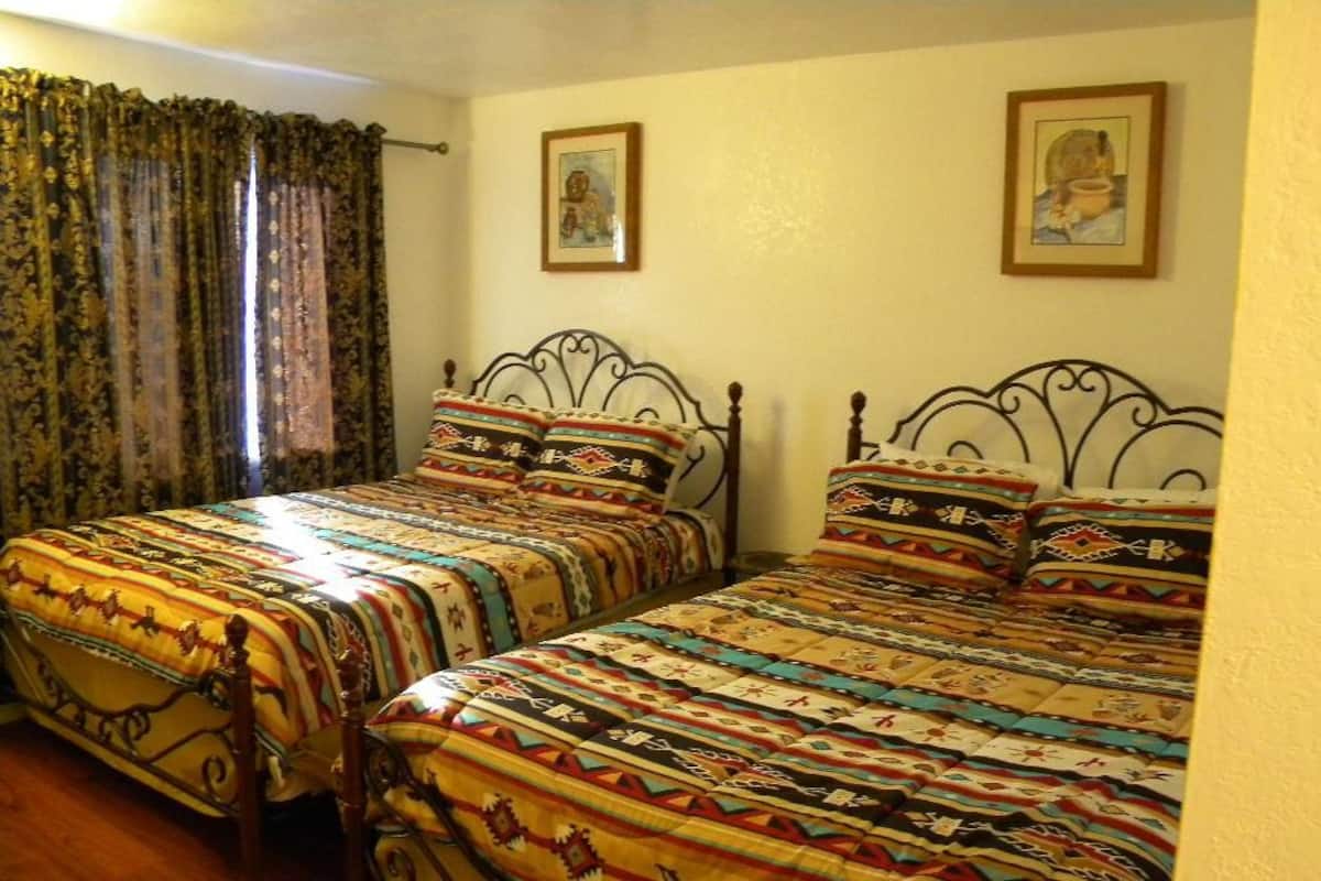 Ein Zimmer mit zwei Betten, jeweils mit einem gemusterten Überwurf und passenden Kissen. Am Fenster sind Vorhänge, und an der Wand hängen zwei gerahmte Bilder.