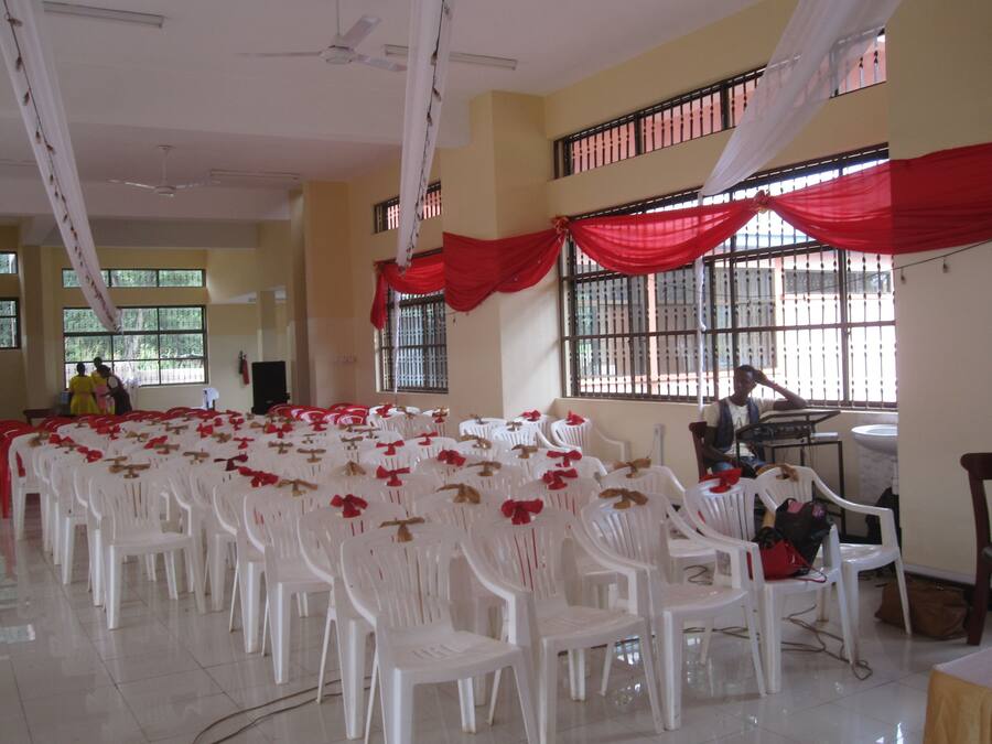 A room with rows of white plastic chairs, a long table, and red drapes.