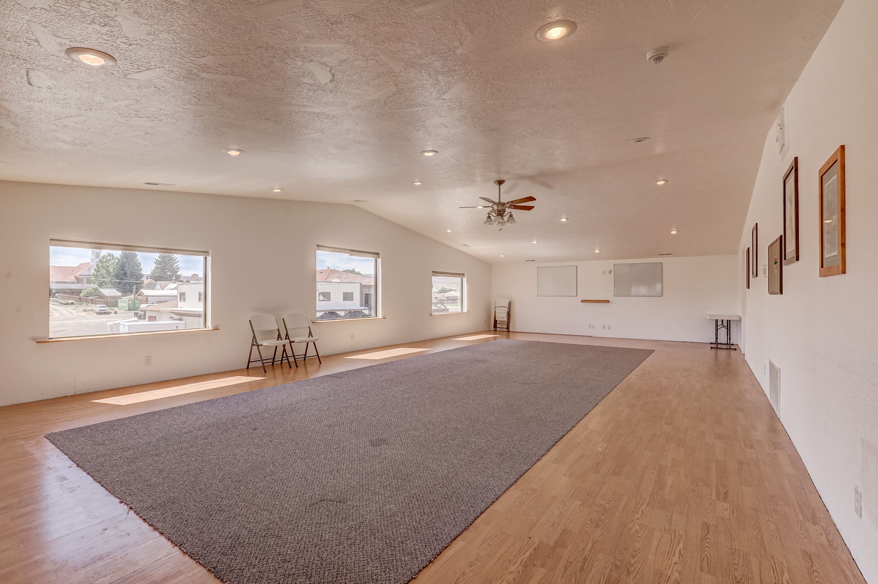 Una habitación amplia con ventanas grandes, un ventilador de techo y alfombra gris.