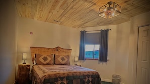 A bedroom with a wooden ceiling, a bed with a patterned bedspread, a nightstand with a lamp, a window with curtains, and a door. - The Snuggle Inn (Loa)