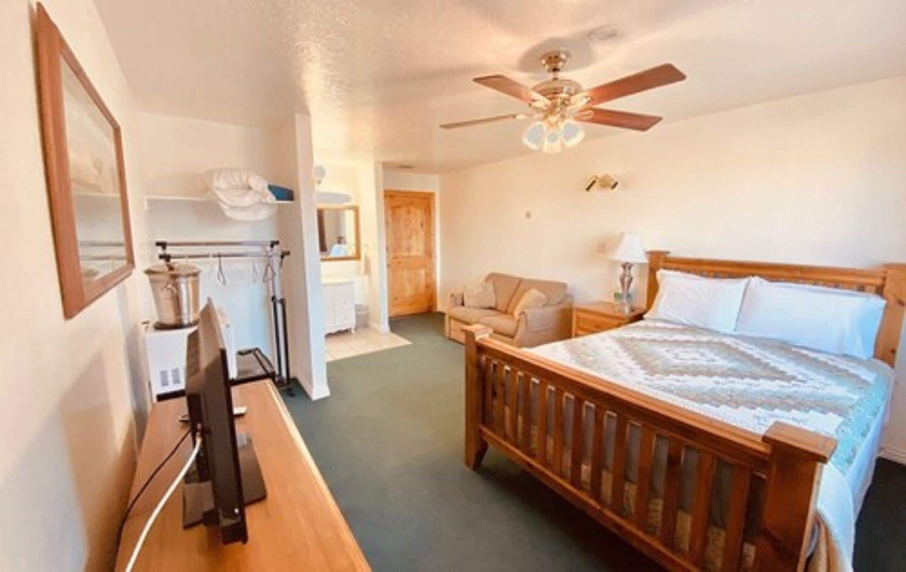 A bedroom with a wooden bed, a desk with a computer, a ceiling fan, and a small seating area with a chair and table.