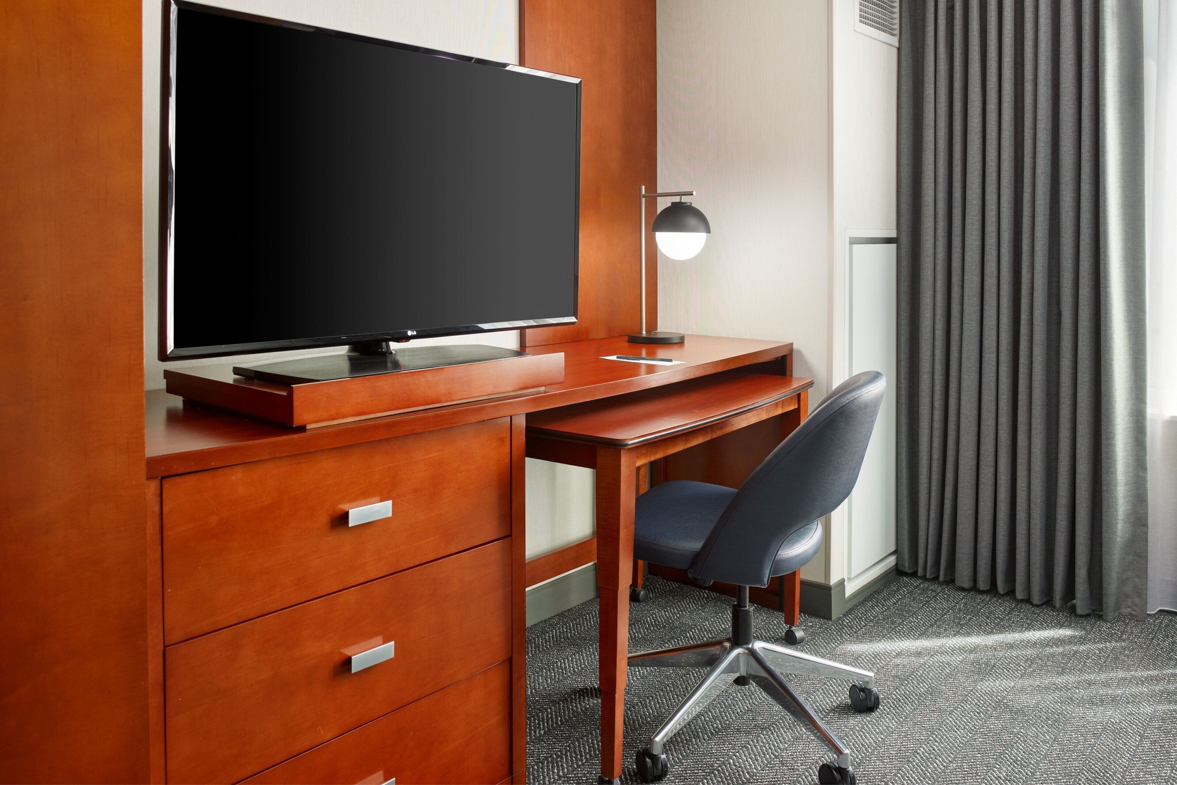 A hotel room with a wooden desk, a flat-screen TV, a modern desk lamp, a black office chair, and a grey carpet.
