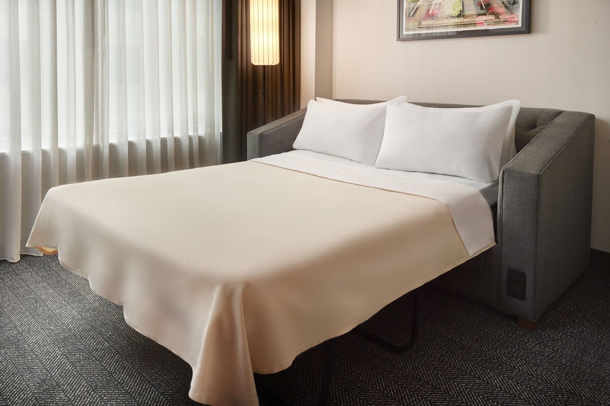A hotel room with a bed, a gray armchair, a window with sheer curtains, a wall-mounted lamp, and a framed picture.
