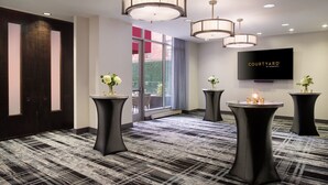 A hotel lobby with a large TV screen displaying "COURTYARD by Marriott", black round tables with vases, a patterned carpet, and a dark wooden door.