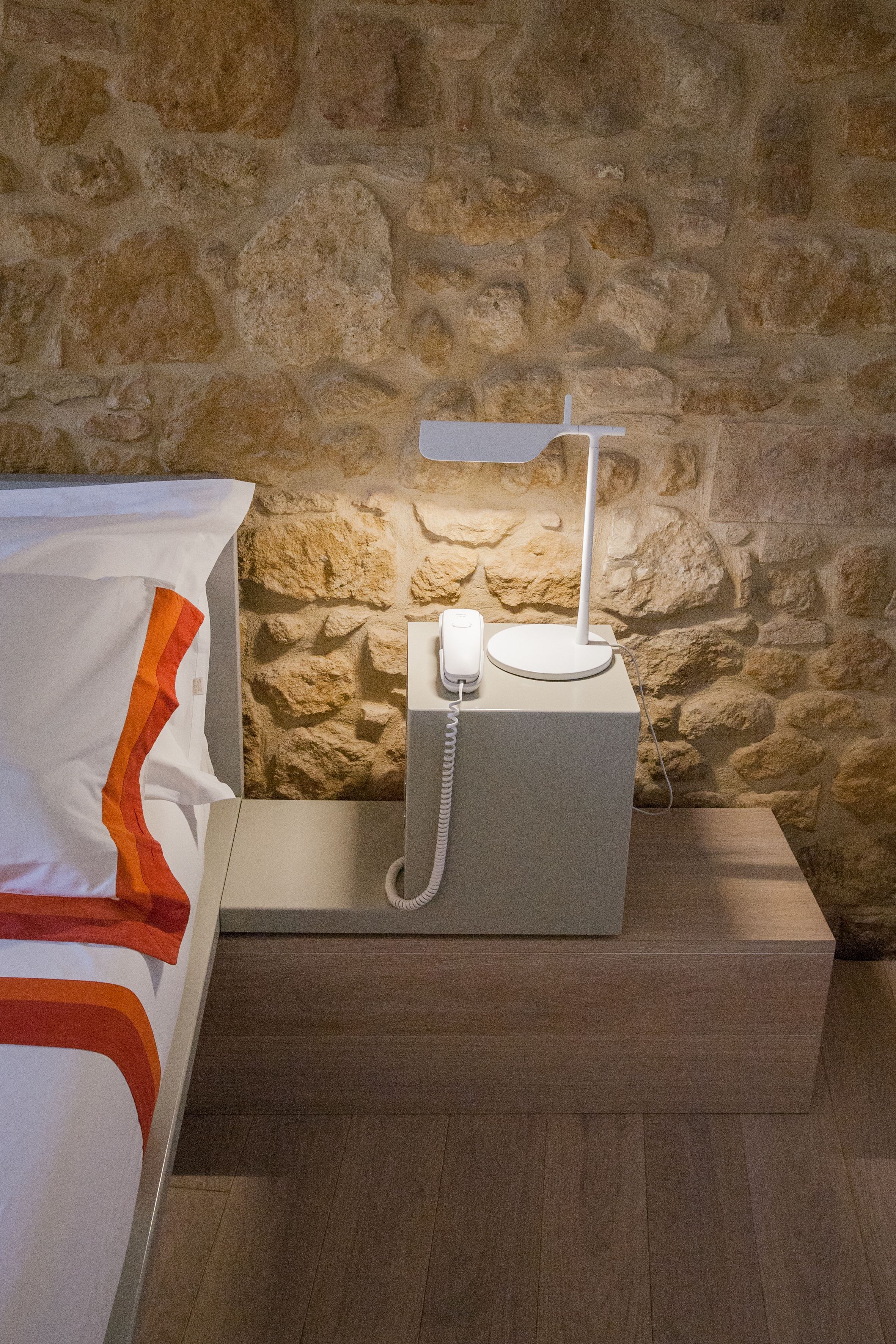 A hotel room with a stone wall, a bedside table with a telephone, and a bed with a white and orange striped blanket.