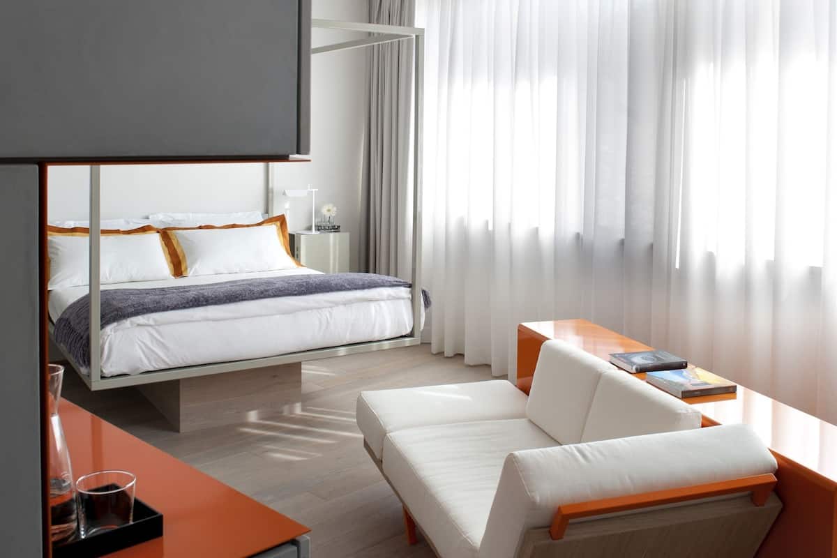 A modern hotel room with a bed, a sofa, a desk, and a large window with curtains.