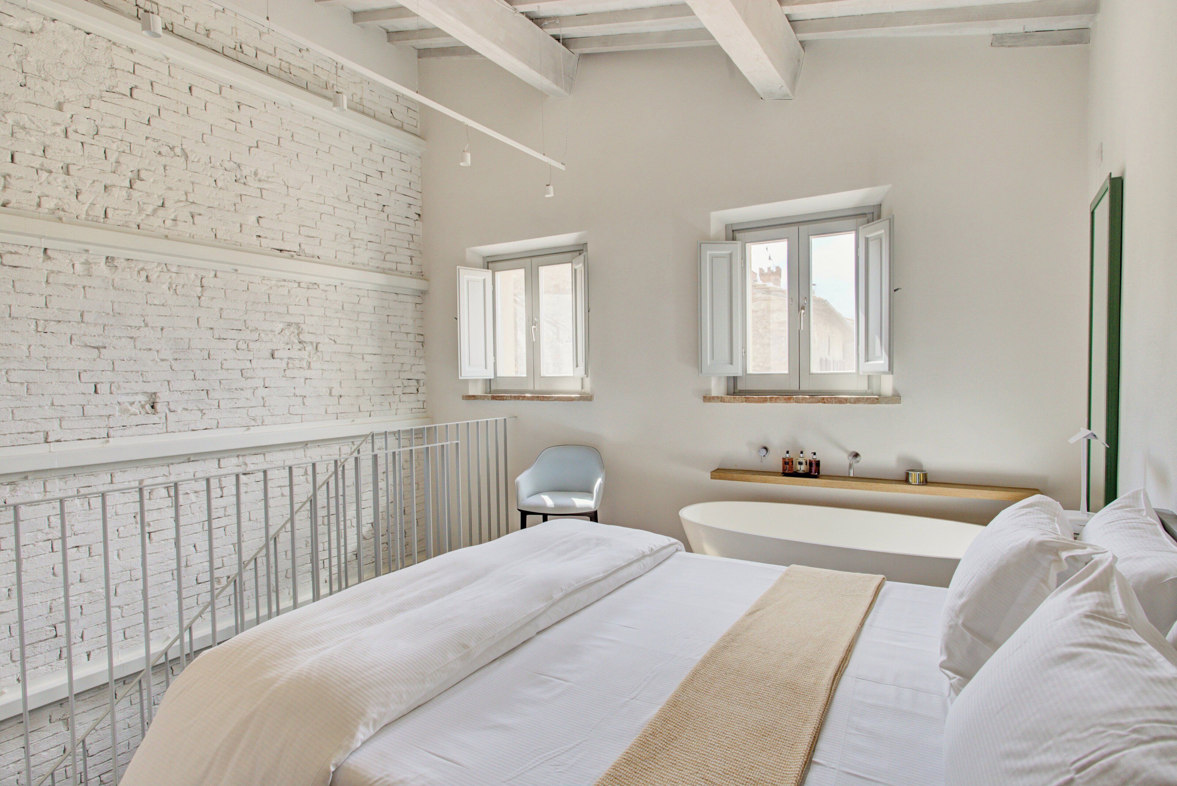 Une chambre avec un mur en briques blanches, un grand lit, une baignoire blanche et deux fenêtres.