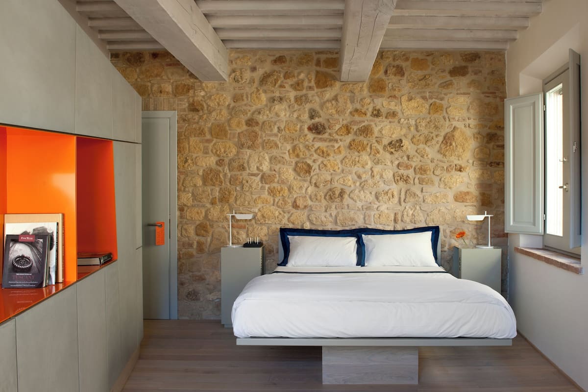 A bedroom with a stone wall, a bed with white bedding, a wooden floor, and a bright orange shelf.