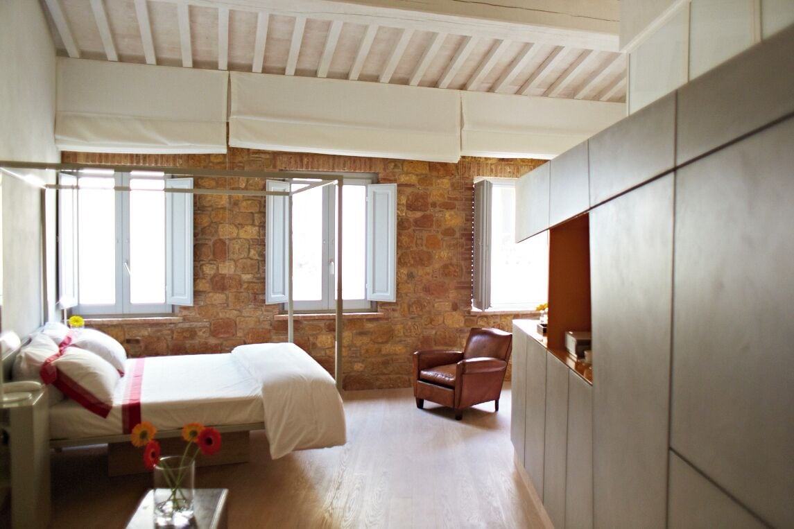 A bedroom with a stone wall, a bed with white linens, a wooden floor, and a window with white shutters.