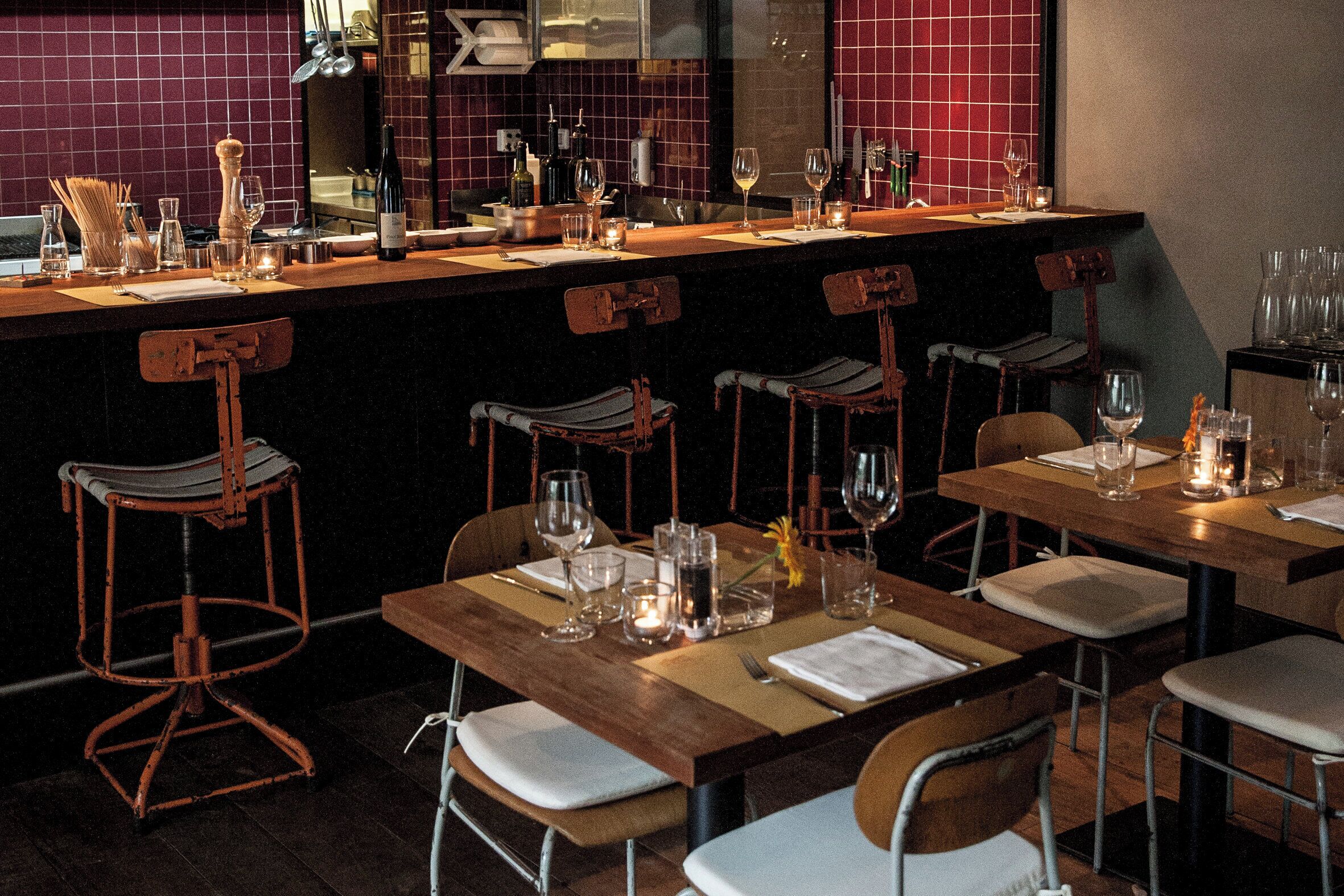 A restaurant interior with wooden tables and chairs, wine glasses, and bottles on the bar.