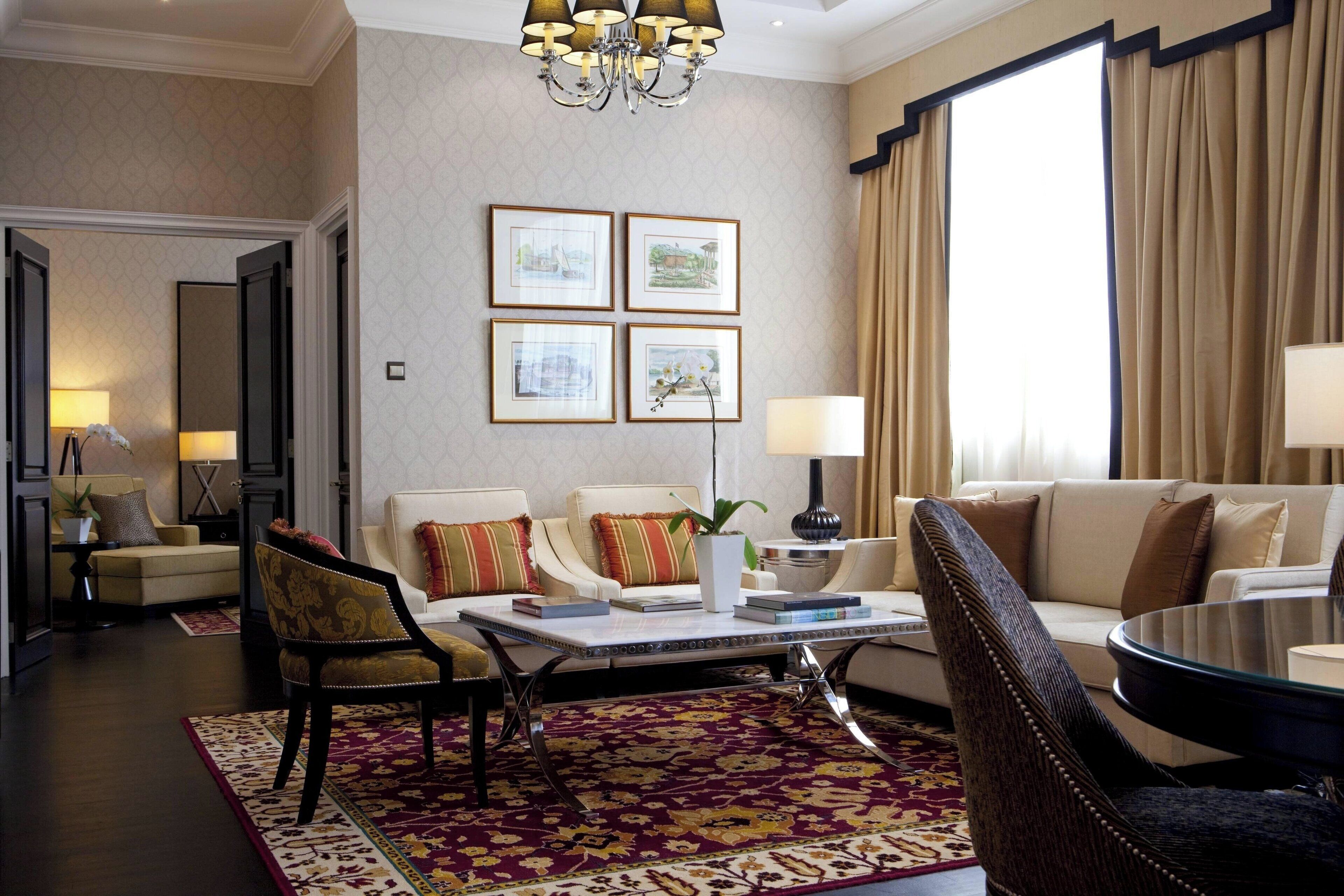 Een goed verlichte woonkamer met een bank, fauteuils, een salontafel en een kroonluchter. Er hangen ingelijste foto’s aan de muur en is een groot raam met gordijnen.