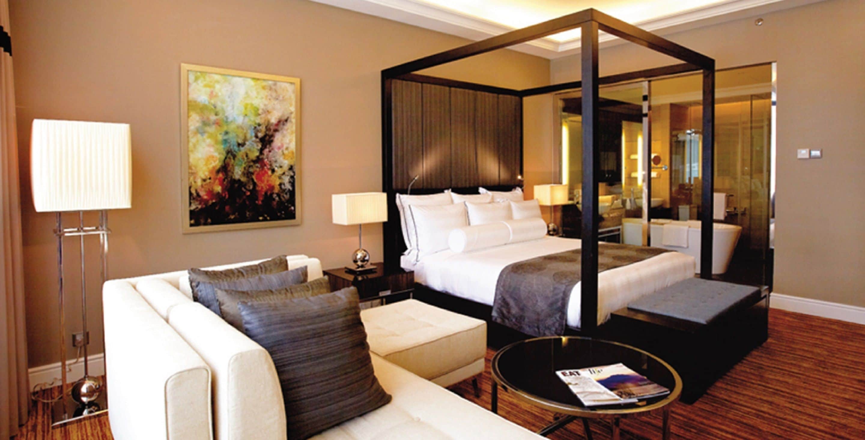 A modern hotel room with a four-poster bed, a sitting area with a sofa and ottoman, a bedside table with a lamp, and a large abstract painting on the wall.