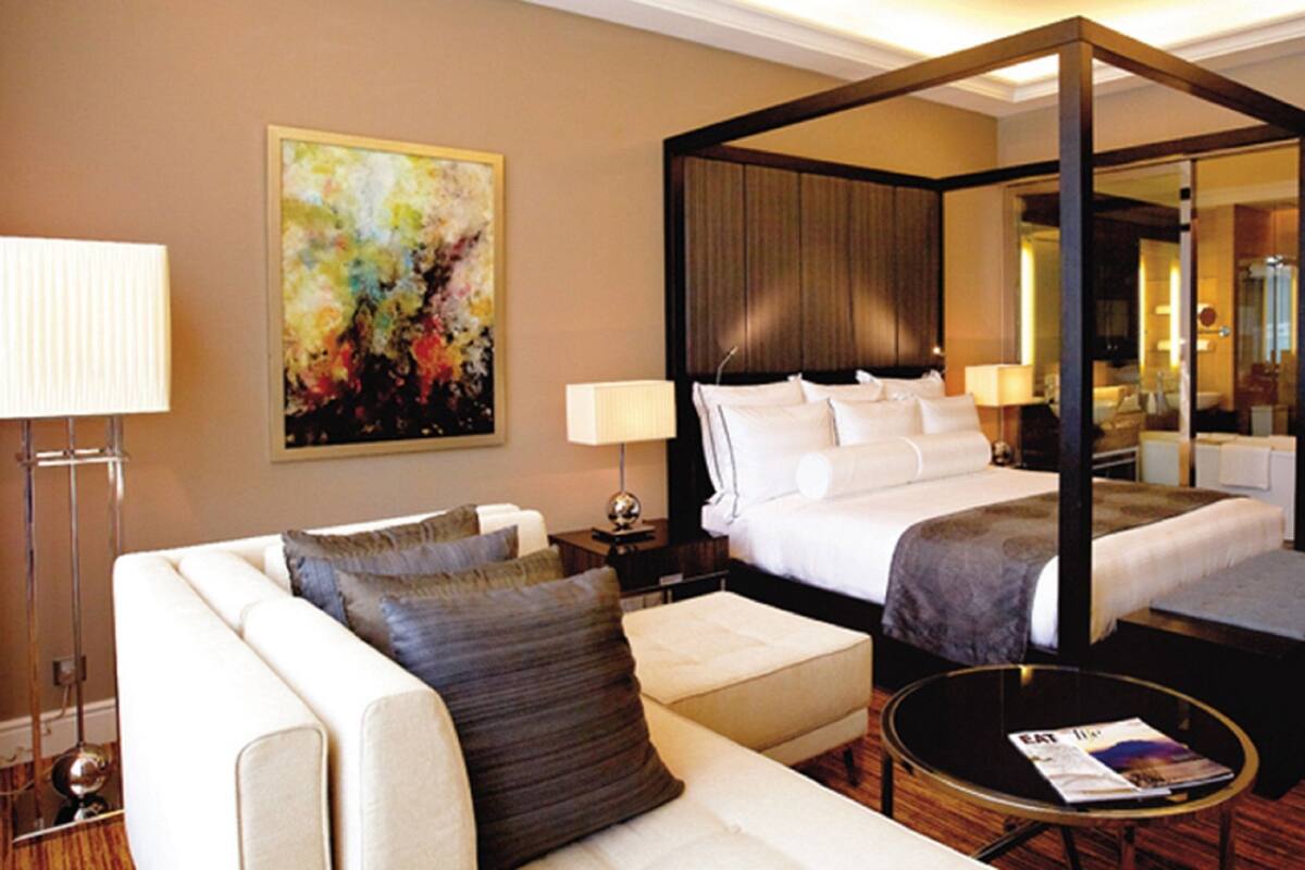 Una habitación de hotel moderna con una cama con dosel, un área de estar con sofá y reposapiés, una mesita de noche con lámpara, y un cuadro abstracto grande en la pared.