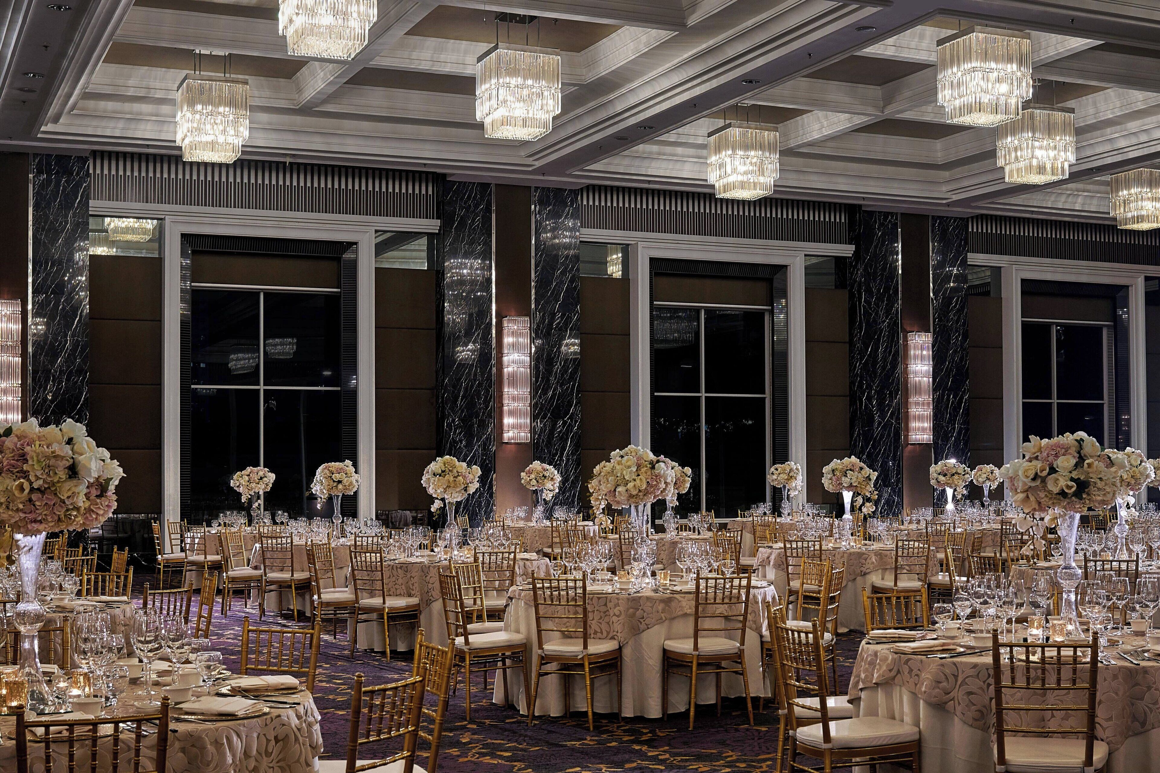 A banquet hall with round tables, gold chairs, and chandeliers.