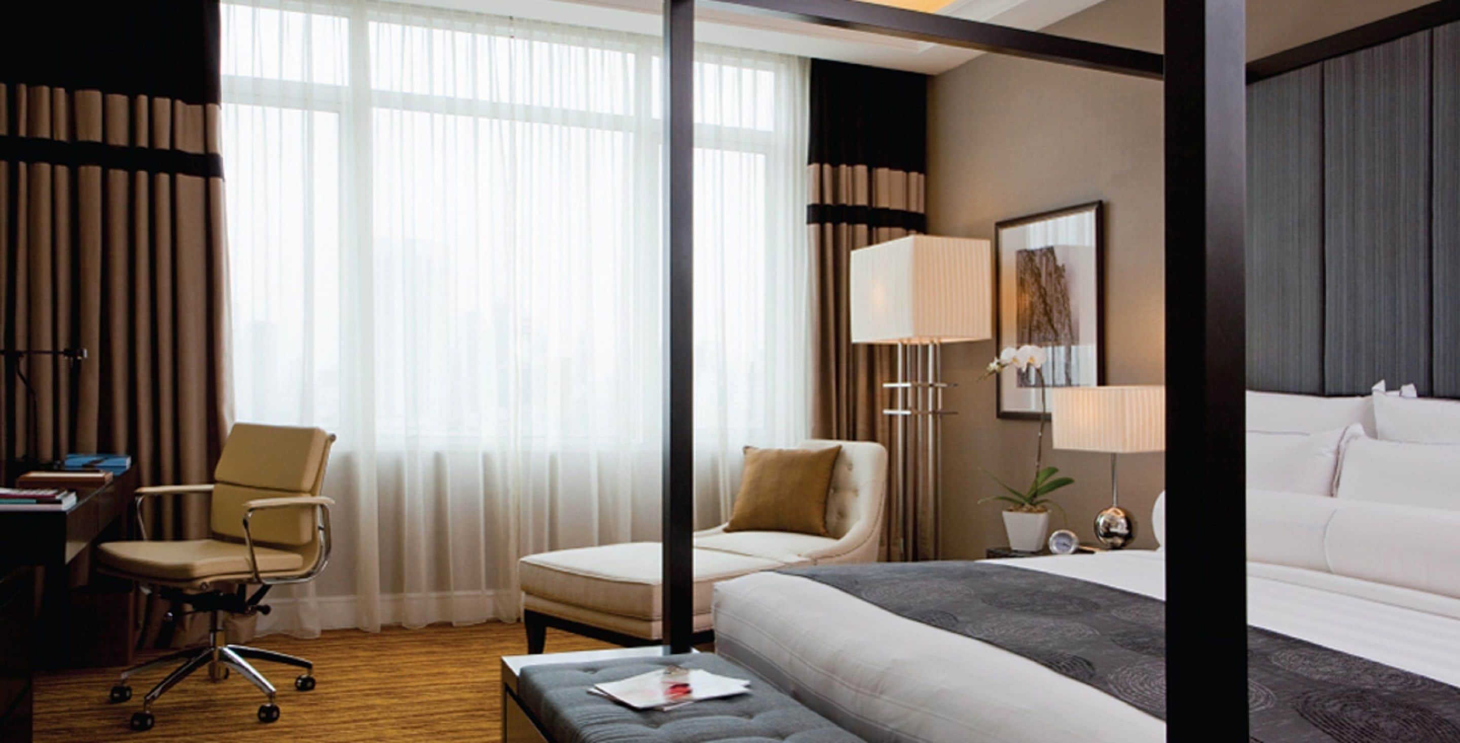 A hotel room with a bed, a desk, a chair, a lamp, and a window with curtains.