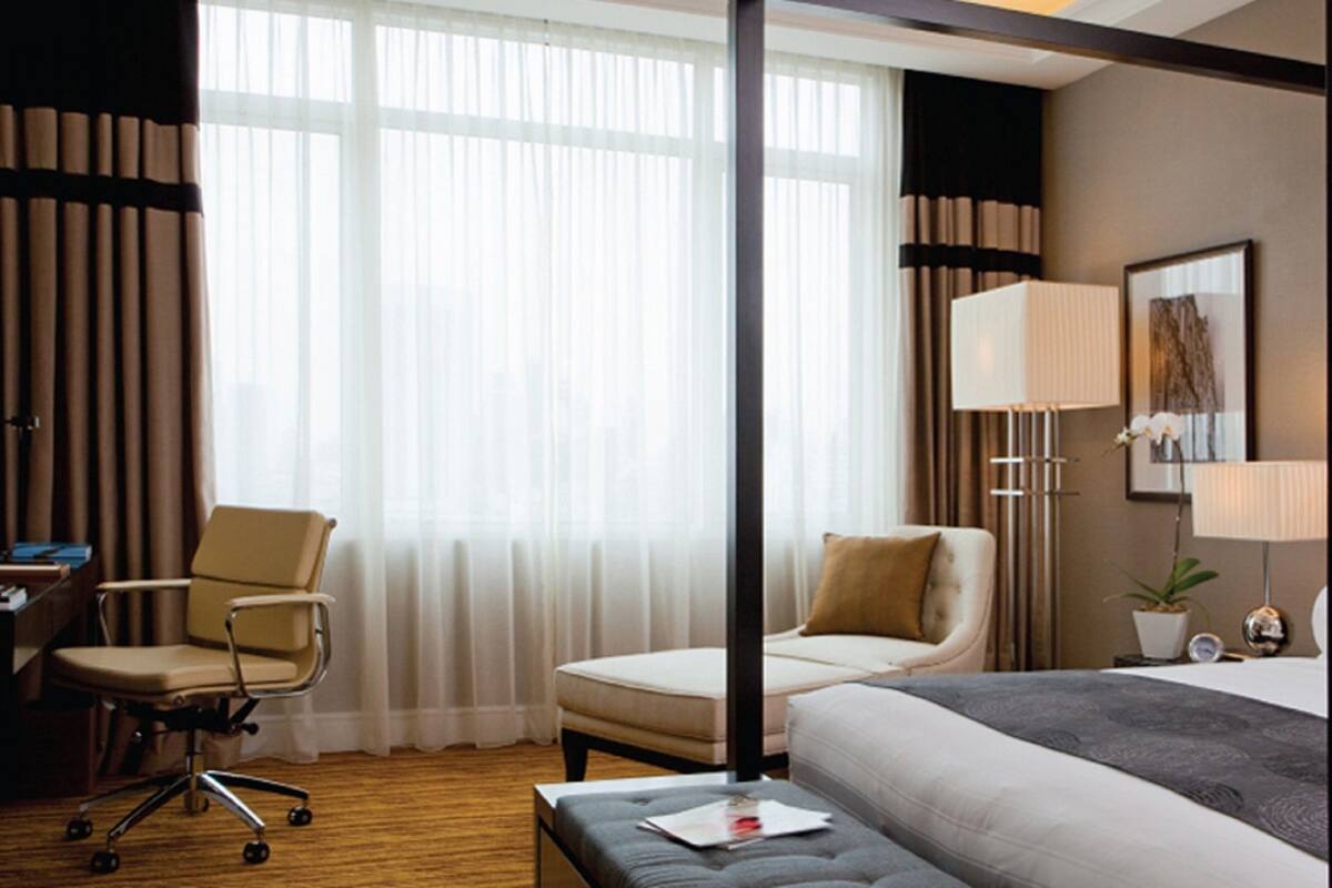 Una habitación de hotel con cama, escritorio, silla, lámpara y ventana con cortinas.