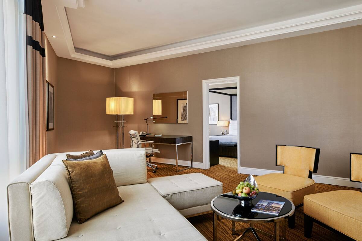 A modern hotel room with a sofa, armchairs, a dining table with fruit, a desk, and a mirror.