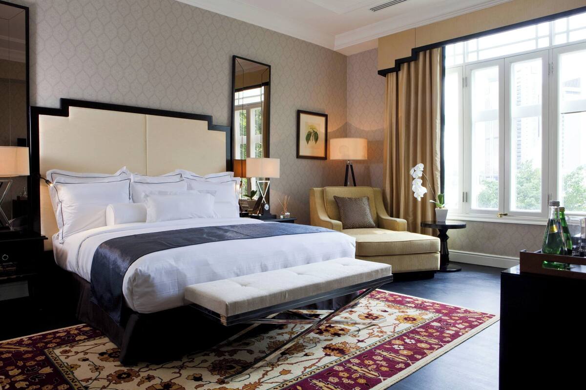 A hotel room with a large bed, a chair, a bench, a desk, a lamp, a mirror, and a window with curtains.