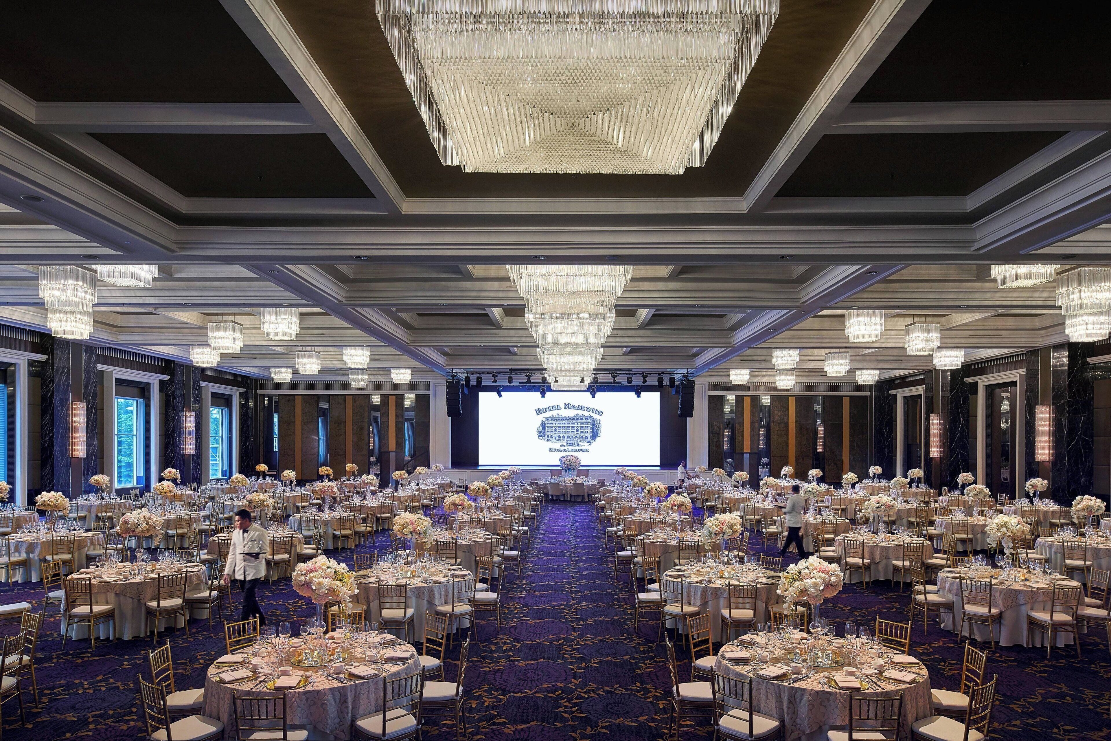 A large banquet hall with round tables, chandeliers, and a large screen at the front.