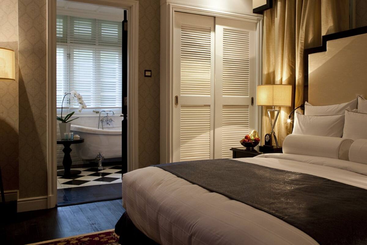 Una habitación de hotel con una cama grande, una mesita de noche con lámpara, un atril y vistas al baño.