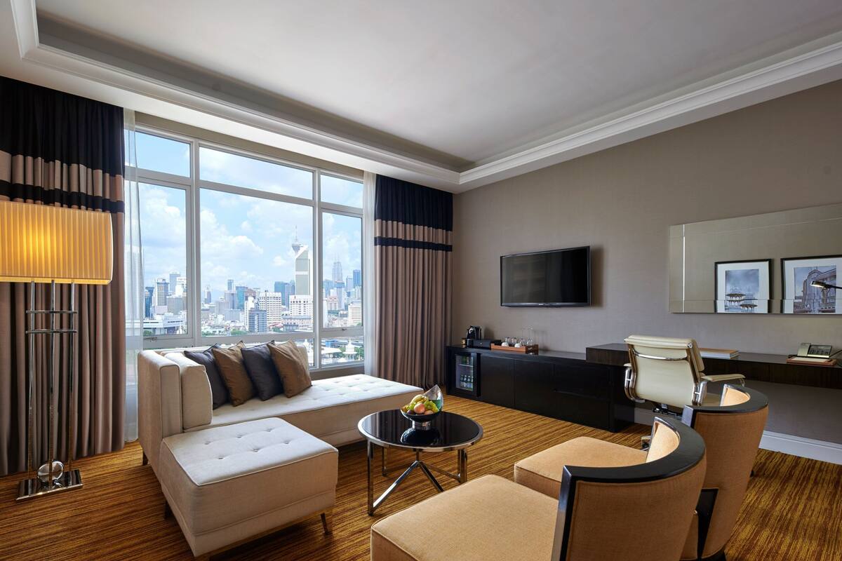 A modern hotel room with a large window, a sofa, a dining table, a TV, and a seating area.