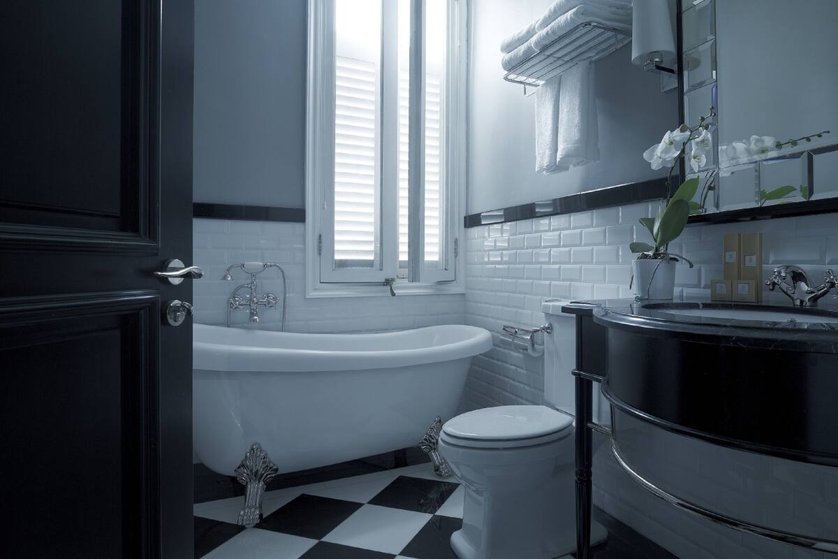 Un baño con piso a cuadros blanco y negro, una bañera blanca, un inodoro y un lavamanos con espejo.