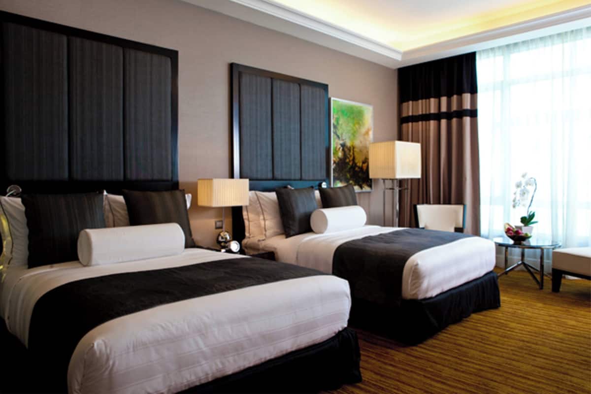 A hotel room with two beds, a nightstand, a lamp, a chair, and a window with curtains.