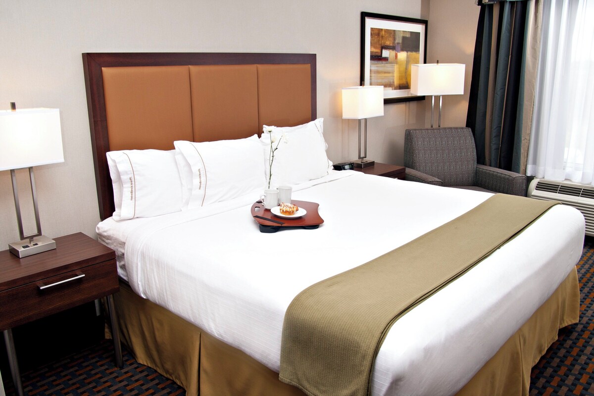 A hotel room with a large bed, a bedside table with a lamp, a chair, and a tray with a plate of food.