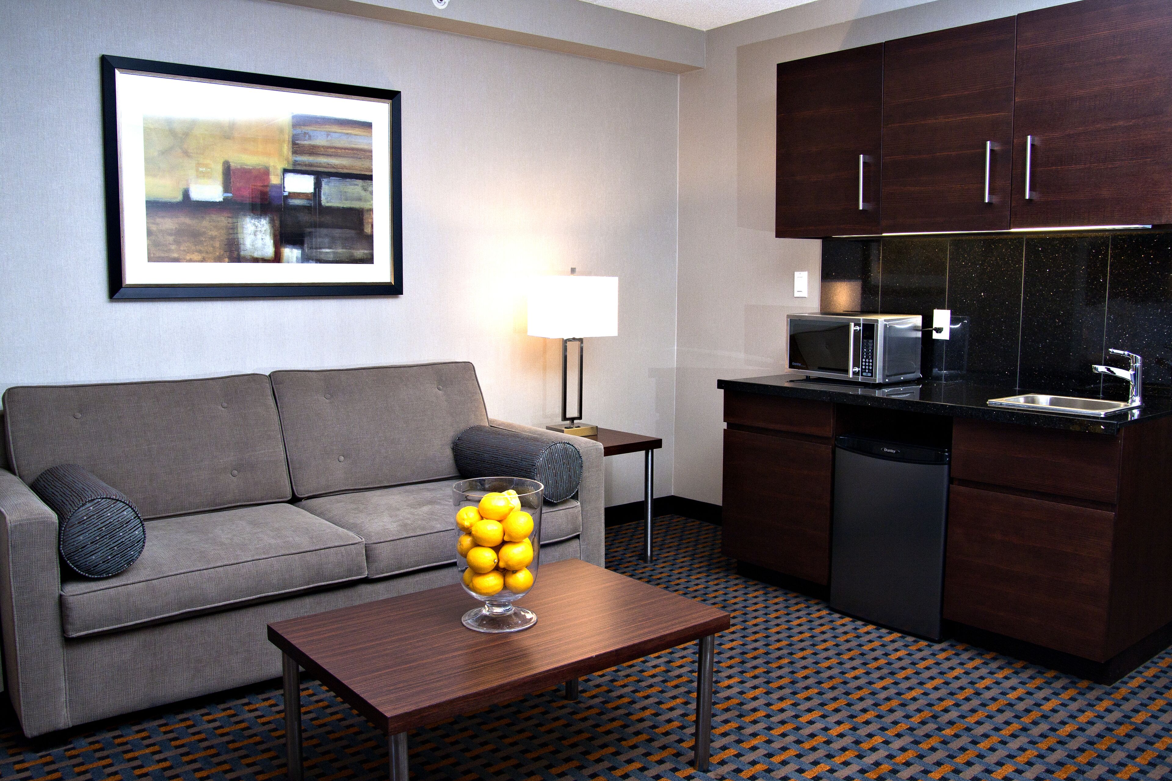 A modern hotel room with a grey sofa, a coffee table with a glass vase of lemons, a kitchen area with a microwave and dishwasher, and a patterned floor.