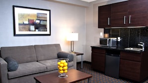 A modern hotel room with a grey sofa, a coffee table with a glass vase of lemons, a kitchen area with a microwave and dishwasher, and a patterned floor. - Holiday Inn Express Pembroke by IHG (Pembroke)