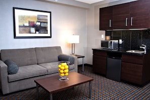 A modern hotel room with a grey sofa, a coffee table with a glass vase of lemons, a kitchen area with a microwave and dishwasher, and a patterned floor. - Holiday Inn Express Pembroke by IHG (Pembroke)