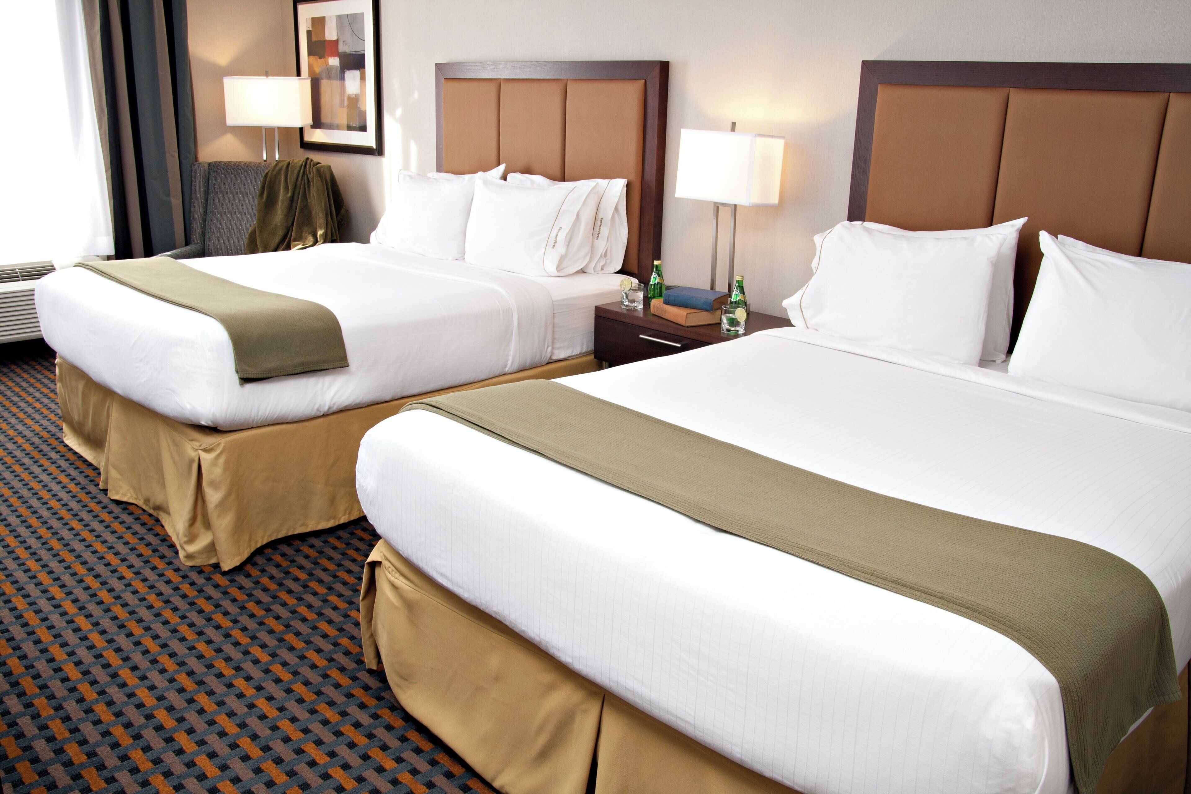 A hotel room with two beds, each with a brown bedspread, white linens, and a headboard. There are two bedside tables with lamps, a framed picture on the wall, and a patterned carpet.