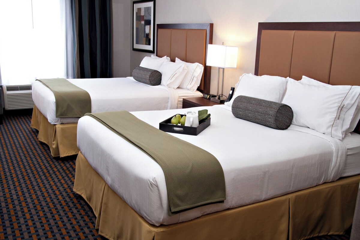 Two hotel beds with white linens, brown headboards, and a tray with fruits and a bottle on the bed.
