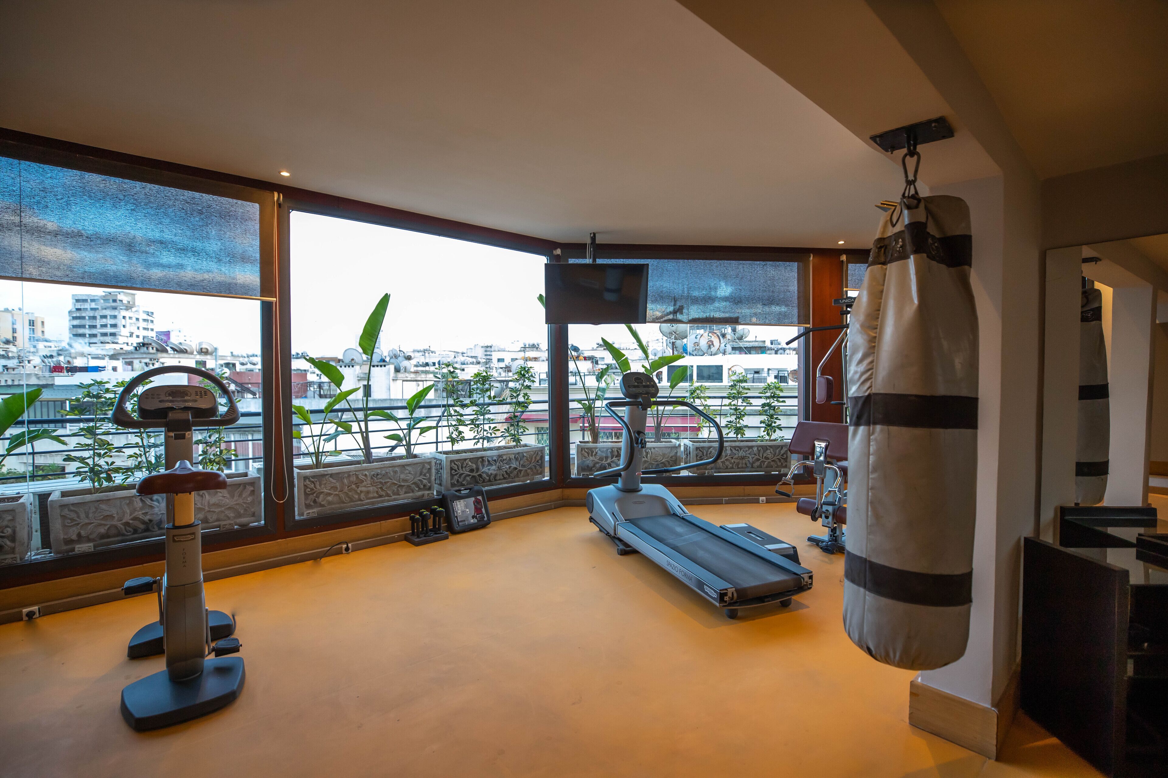 Ein Fitnessraum mit Laufband, Ergometer und Sandsack, mit Blick auf eine Stadtlandschaft durch große Fenster.
