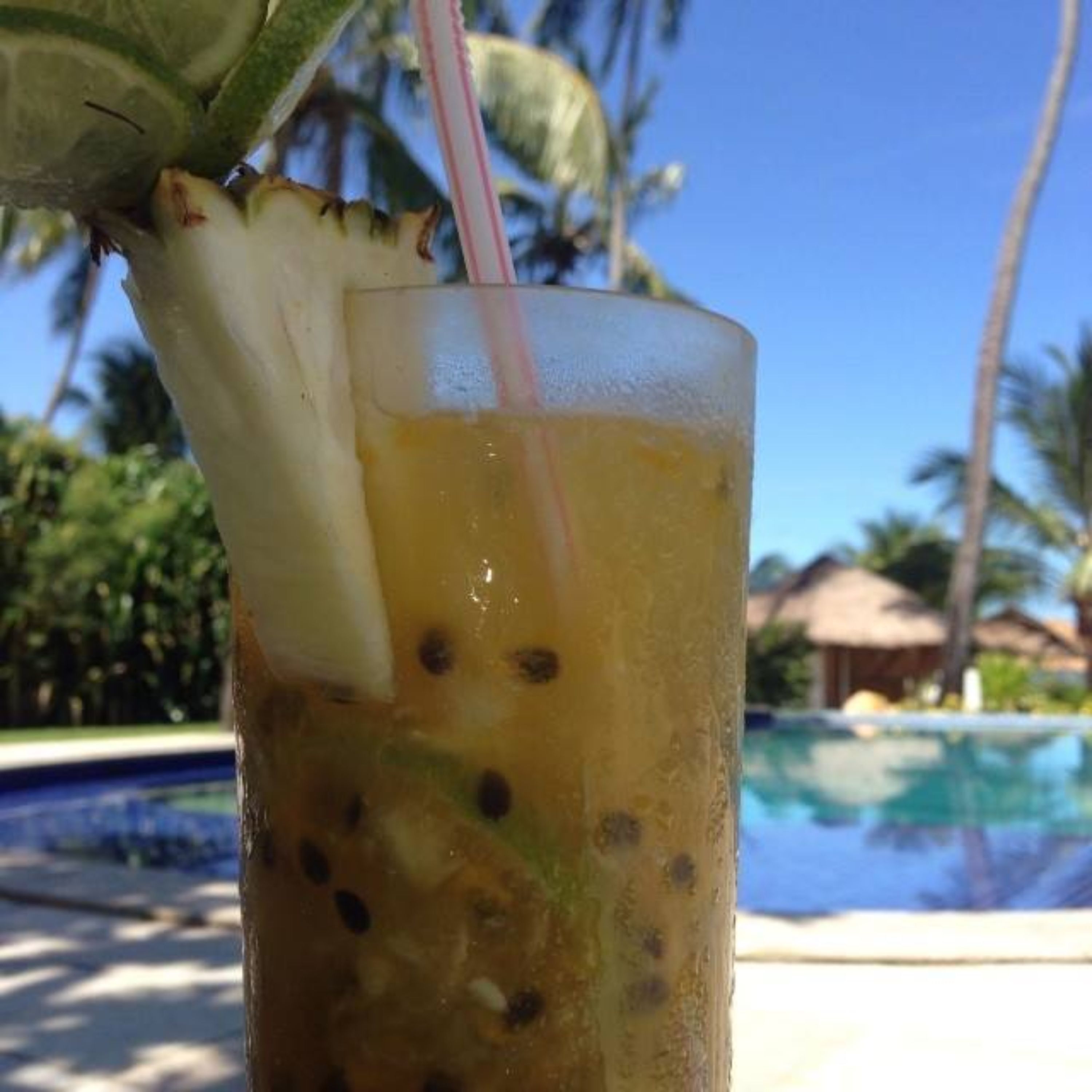 A glass with a straw and a slice of fruit, likely a tropical drink, near a pool with palm trees and a thatched-roof structure in the background.