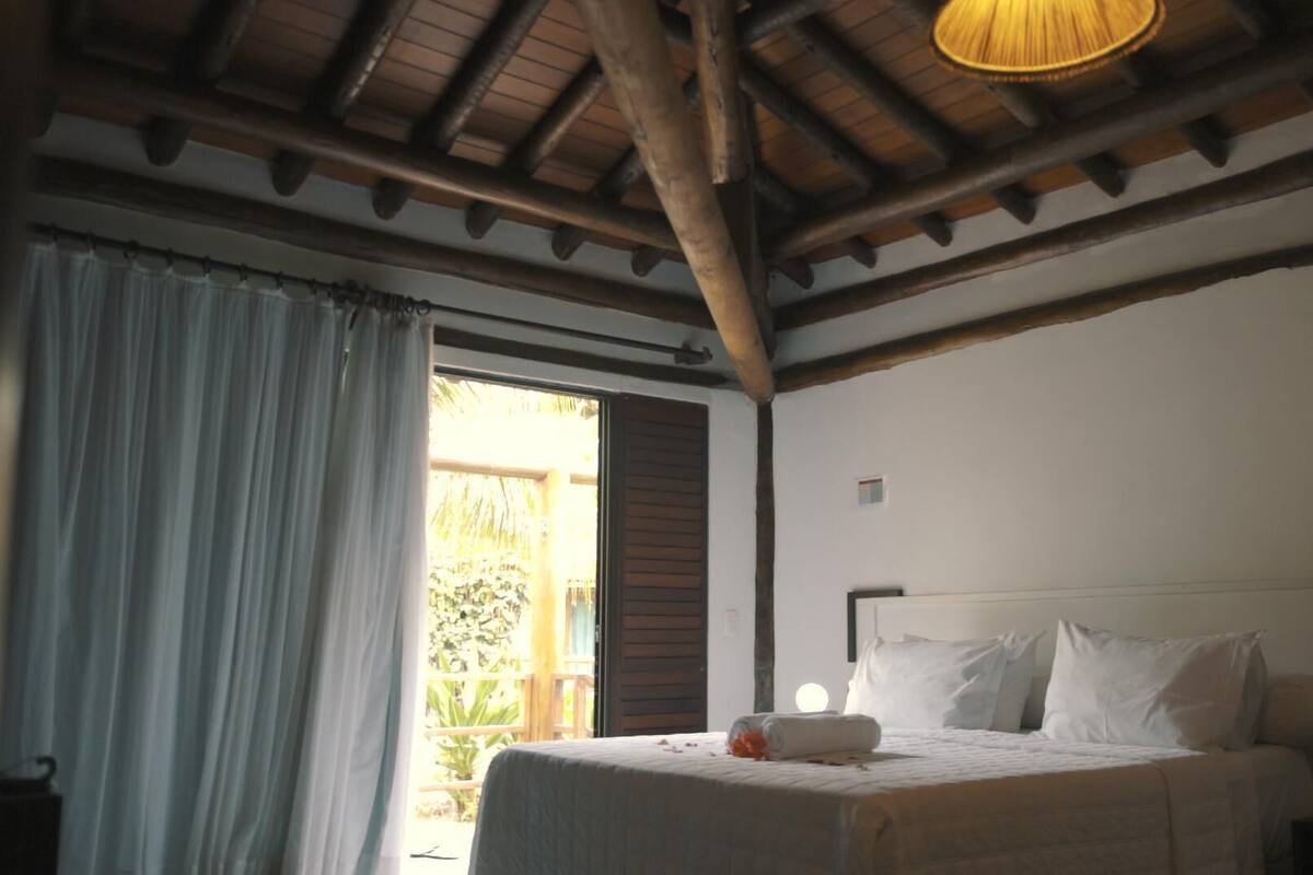 Un dormitorio con una cama, una ventana con cortinas, techo de madera y una mesita.