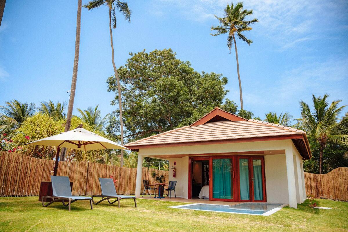 Una pequeña casa con techo de tejas, un porche cubierto con sillas y una mesa, y una piscina.