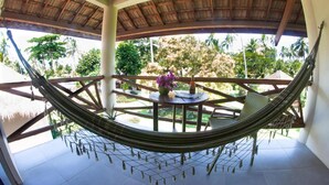 A hammock chair under a wooden roof, a table with a vase of flowers, and a view of greenery outside.