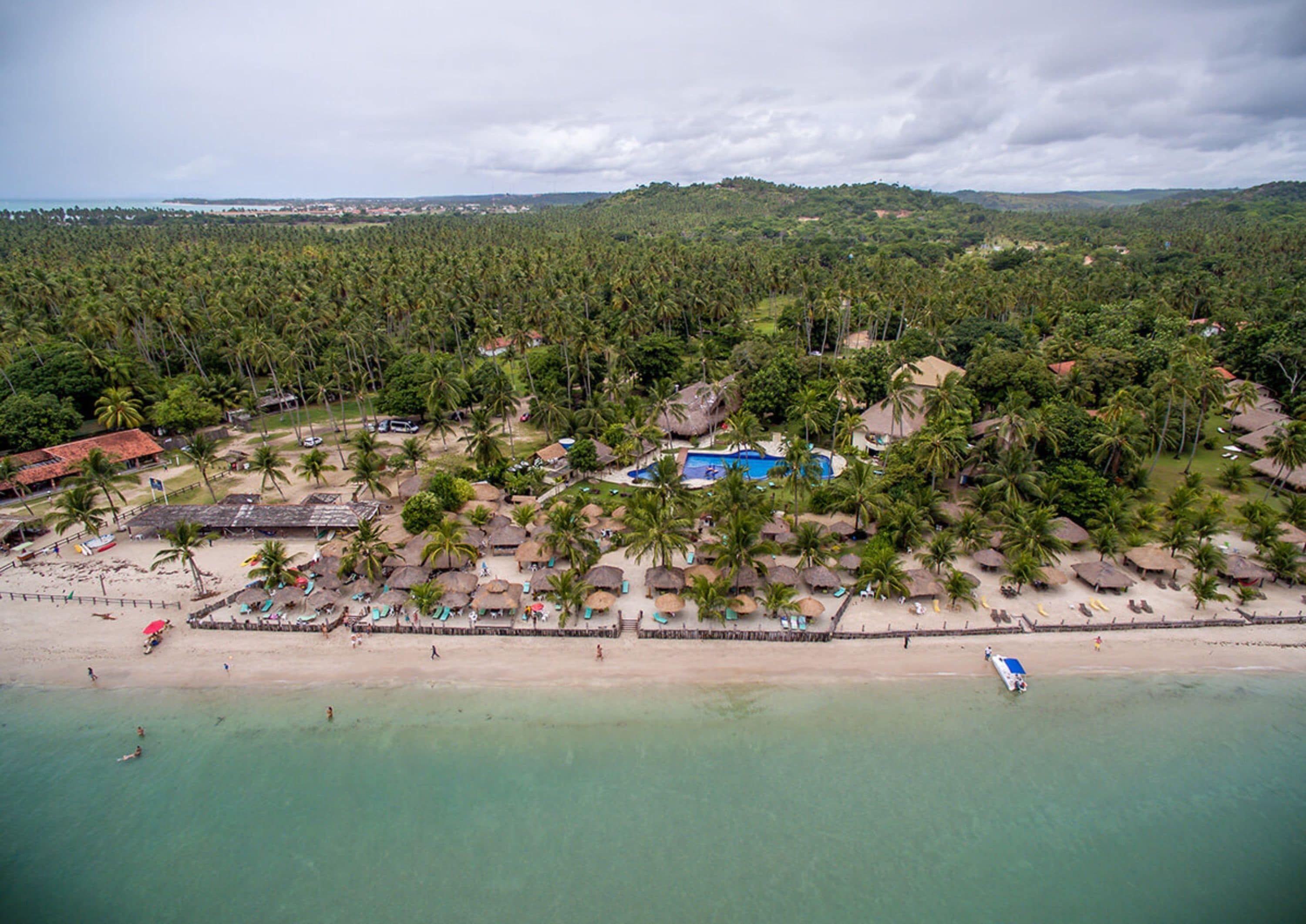 An aerial view of a beach resort with a swimming pool, palm trees, and beach umbrellas.